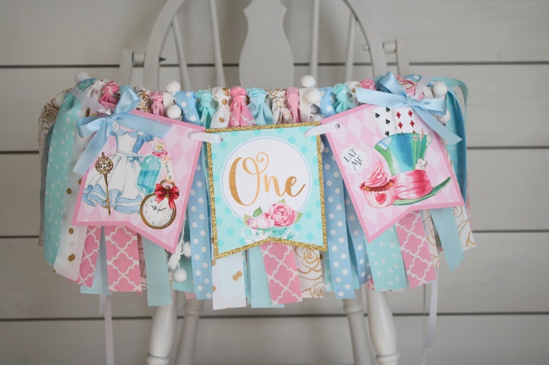 May include: A pink and blue Alice in Wonderland themed birthday banner with the word "One" in gold glitter. The banner features images of Alice, the Mad Hatter, and a cupcake.
