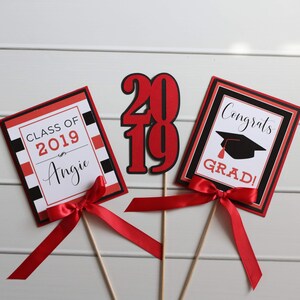 May include: Three graduation party decorations with red and black accents. The first decoration is a red and black card with the text "Class of 2019 Angie". The second decoration is a red and black card with the text "Congrats Grad!" and a black graduation cap. The third decoration is a red and black "2019" cutout.