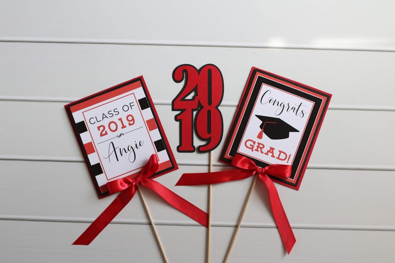 May include: Three graduation party decorations with red and black accents. The first decoration is a red and black card with the text "Class of 2019 Angie". The second decoration is a red and black card with the text "Congrats Grad!" and a black graduation cap. The third decoration is a red and black "2019" cutout.