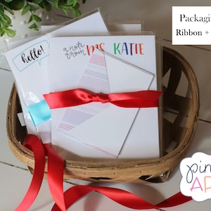 Puede incluir: Un conjunto de tres tarjetas de notas en blanco con diferentes dise&ntilde;os, que incluyen "hello!" en un globo de di&aacute;logo, "a note from" con un dise&ntilde;o de arco&iacute;ris y "Mrs. Katie" en letras de arco&iacute;ris. Las tarjetas est&aacute;n envueltas en una cesta con una cinta roja y un mensaje de regalo.