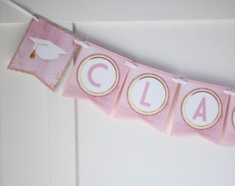 May include: Pink and gold graduation banner with the word "CLASS" in white letters on each flag. The flags are decorated with gold glitter and a white graduation cap.
