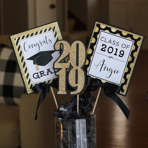 Puede incluir: Centro de mesa para una fiesta de graduaci&oacute;n con decoraciones negras y doradas. El centro de mesa incluye un letrero que dice "Congrats Grad", un letrero que dice "Class of 2019 Angie", y un letrero grande de purpurina dorada "2019".