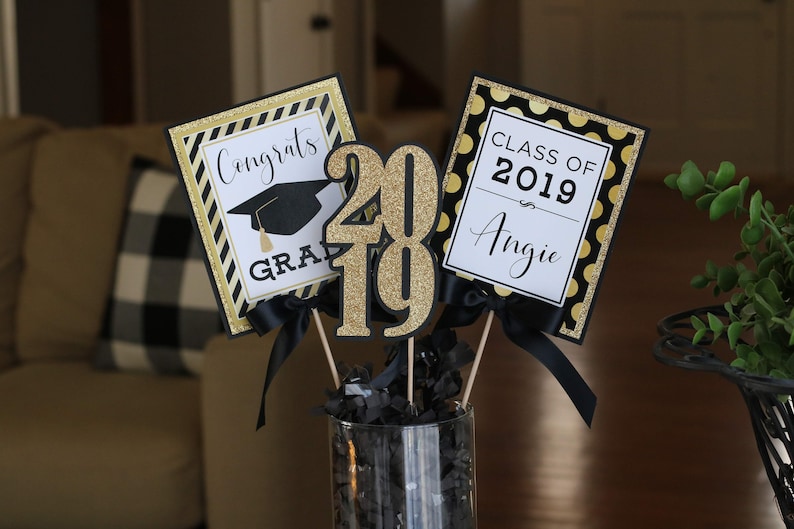 Puede incluir: Centro de mesa para una fiesta de graduaci&oacute;n con decoraciones negras y doradas. El centro de mesa incluye un letrero que dice "Congrats Grad", un letrero que dice "Class of 2019 Angie", y un letrero grande de purpurina dorada "2019".