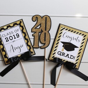 Puede incluir: Tres decoraciones para una fiesta de graduaci&oacute;n con detalles de purpurina negra y dorada. La primera decoraci&oacute;n dice "Clase de 2019 Angie". La segunda decoraci&oacute;n es un "2019" de purpurina dorada. La tercera decoraci&oacute;n dice "&iexcl;Felicidades graduado!" con un gorro de graduaci&oacute;n negro.