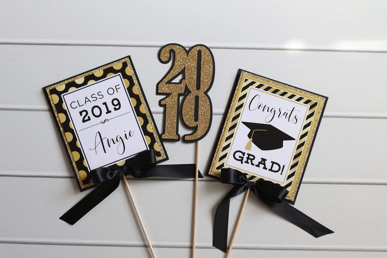 Puede incluir: Tres decoraciones para una fiesta de graduaci&oacute;n con detalles de purpurina negra y dorada. La primera decoraci&oacute;n dice "Clase de 2019 Angie". La segunda decoraci&oacute;n es un "2019" de purpurina dorada. La tercera decoraci&oacute;n dice "&iexcl;Felicidades graduado!" con un gorro de graduaci&oacute;n negro.