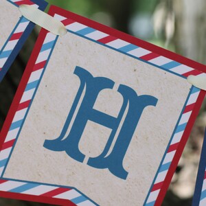 May include: A red, white, and blue striped banner with the letter "H" printed in blue. The banner is made of paper and has a rustic look.
