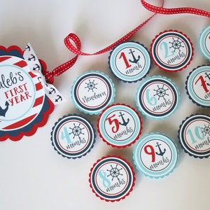 Może przedstawiać: Baner z kamieniami milowymi dla dziecka w stylu marynistycznym z czerwonymi i niebieskimi paskami, kotwicą i tekstem "Caleb's First Year". Baner jest ozdobiony 12 okrągłymi, falistymi k&oacute;łkami, z kt&oacute;rych każde zawiera inną liczbę od 1 do 12, kotwicę lub koło sterowe, a także słowo "miesiące".