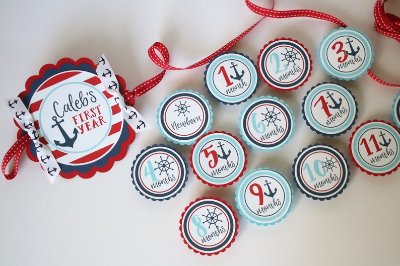 Może przedstawiać: Baner z kamieniami milowymi dla dziecka w stylu marynistycznym z czerwonymi i niebieskimi paskami, kotwicą i tekstem "Caleb's First Year". Baner jest ozdobiony 12 okrągłymi, falistymi k&oacute;łkami, z kt&oacute;rych każde zawiera inną liczbę od 1 do 12, kotwicę lub koło sterowe, a także słowo "miesiące".