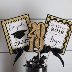 Puede incluir: Centro de mesa de graduaci&oacute;n negro y dorado con un gorro de graduaci&oacute;n negro, las palabras "Congrats Grad" y "Class of 2019 Angie" y el n&uacute;mero "2019" en purpurina dorada.