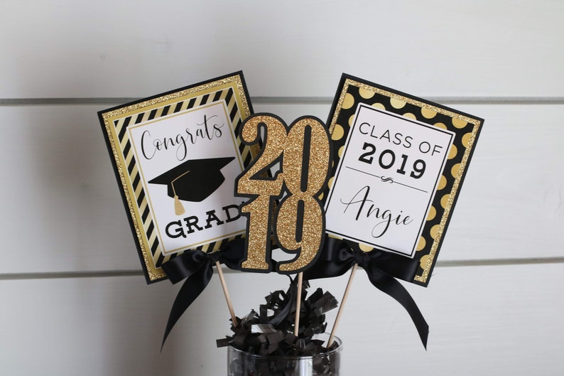 Puede incluir: Centro de mesa de graduaci&oacute;n negro y dorado con un gorro de graduaci&oacute;n negro, las palabras "Congrats Grad" y "Class of 2019 Angie" y el n&uacute;mero "2019" en purpurina dorada.