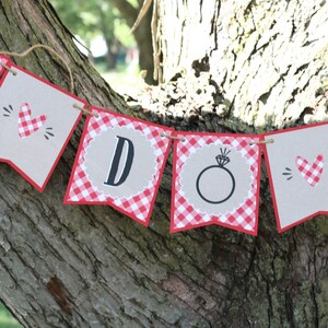 May include: A red and white gingham banner with the words "I Do" in black letters. The banner is decorated with hearts and a ring.