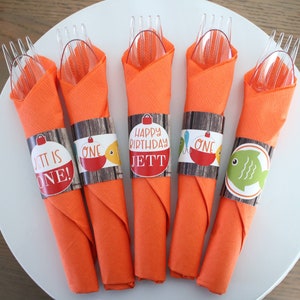 May include: Orange napkins with clear plastic forks and spoons are arranged on a white plate. Each napkin is wrapped with a decorative band featuring a fishing theme and text that reads "It is ONE!" and "Happy Birthday Jett".