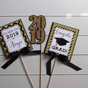 Puede incluir: Tres decoraciones para una fiesta de graduaci&oacute;n con acentos negros y dorados. Un letrero dice "Clase de 2019 Angie", otro dice "&iexcl;Felicidades graduado!" y el tercero es un gran "2019" de purpurina dorada.