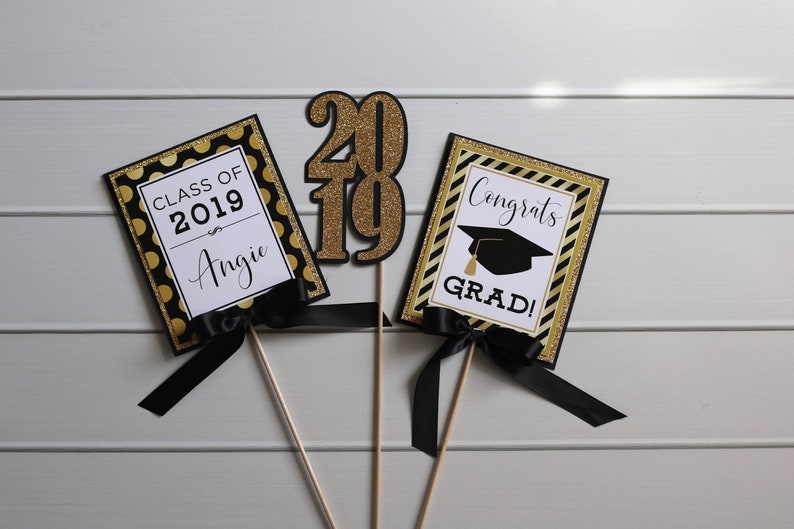 Puede incluir: Tres decoraciones para una fiesta de graduaci&oacute;n con acentos negros y dorados. Un letrero dice "Clase de 2019 Angie", otro dice "&iexcl;Felicidades graduado!" y el tercero es un gran "2019" de purpurina dorada.