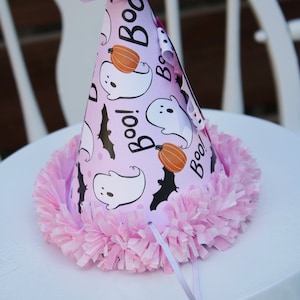 May include: A pink Halloween party hat with a ruffled trim and a paper flower on top. The hat features white ghosts, orange pumpkins, and black bats on a light purple background, with the word "Boo!" printed on it.