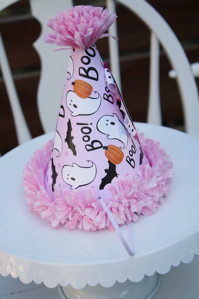 May include: A pink Halloween party hat with a ruffled trim and a paper flower on top. The hat features white ghosts, orange pumpkins, and black bats on a light purple background, with the word "Boo!" printed on it.