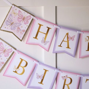 May include: A pink and purple butterfly themed banner with gold letters spelling "Happy Birthday".