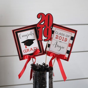 May include: Graduation centerpiece with red and black decorations. The centerpiece includes a sign that says "Congrats Grad!" with a graduation cap, a sign that says "Class of 2019 Angie", and a large red number "2019".
