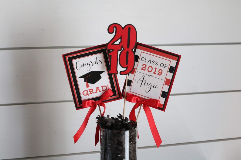 May include: Graduation centerpiece with red and black decorations. The centerpiece includes a sign that says "Congrats Grad!" with a graduation cap, a sign that says "Class of 2019 Angie", and a large red number "2019".