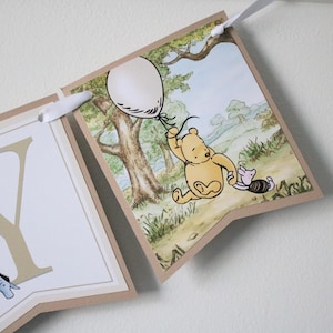May include: A Winnie the Pooh themed banner with a brown background and white letters. The banner features a scene of Winnie the Pooh and Piglet holding balloons. The letters on the banner spell out the word "YOU".
