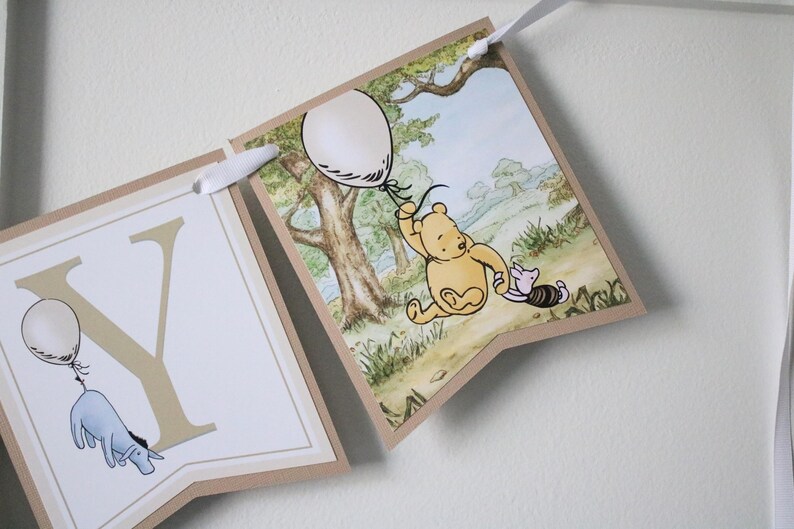 May include: A Winnie the Pooh themed banner with a brown background and white letters. The banner features a scene of Winnie the Pooh and Piglet holding balloons. The letters on the banner spell out the word "YOU".