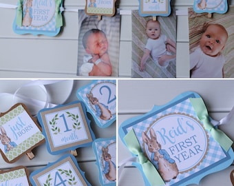 May include: A blue and green Peter Rabbit themed baby milestone banner with a white ribbon. The banner includes a 'Just Born' card, a '1 Month' card, a '2 Months' card, a '3 Months' card, a '4 Months' card, and a 'Reid's First Year' card. Each card features a Peter Rabbit illustration and a number.