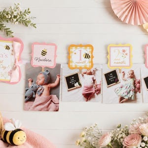 Puede incluir: Una guirnalda de hitos para bebés con un cartel que dice Willa's First Bee-Day, seguido de cuatro tarjetas fotográficas etiquetadas como Newborn, 1 month, 2 months y 3 months. Las tarjetas están decoradas con ilustraciones de abejas y bordes rosas.