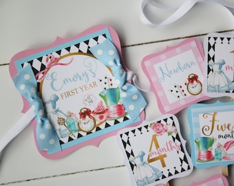 May include: A blue and pink Alice in Wonderland themed baby milestone banner with a white ribbon. The banner includes a title card that says "Emory's First Year" and cards for each month of the baby's first year. The cards feature illustrations of Alice in Wonderland characters and objects.