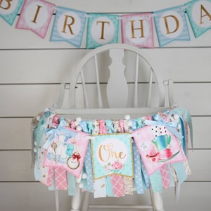 May include: A white high chair with a colorful Alice in Wonderland themed banner draped across the front. The banner features images of Alice, the Mad Hatter, and a teacup. The banner also includes the words "Happy Birthday" and "One".