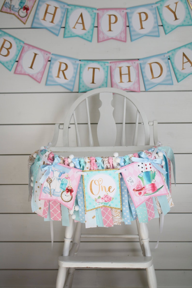 May include: A white high chair with a colorful Alice in Wonderland themed banner draped across the front. The banner features images of Alice, the Mad Hatter, and a teacup. The banner also includes the words "Happy Birthday" and "One".