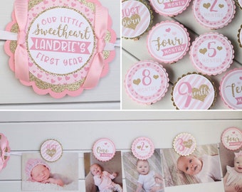 May include: Pink and gold glitter baby milestone banner with a pink ribbon and the text "Our Little Sweetheart Landrie's First Year". The banner includes individual circles with numbers 1 through 9 and the text "Just Born", "One Month", "Two Months", "Four Months", "Five Months", "Eight Months", and "Nine Months".