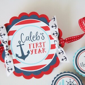 Może przedstawiać: Baner w marynistycznym stylu w kolorze czerwonym, białym i niebieskim z napisem "Caleb's First Year" i kotwicą. Baner jest przewleczony przez białą wstążkę z nadrukowanymi kotwicami i czerwoną wstążkę w białe kropki.