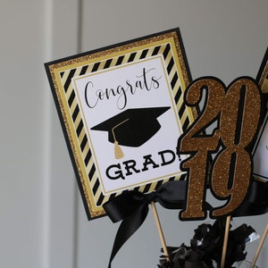 Puede incluir: Decoraciones para una fiesta de graduaci&oacute;n con acentos negros y dorados. Las decoraciones incluyen un letrero que dice "Congrats Grad!" con un gorro de graduaci&oacute;n, un letrero que dice "Class of 2019 Angie", y un gran centro de mesa de purpurina dorada "2019".