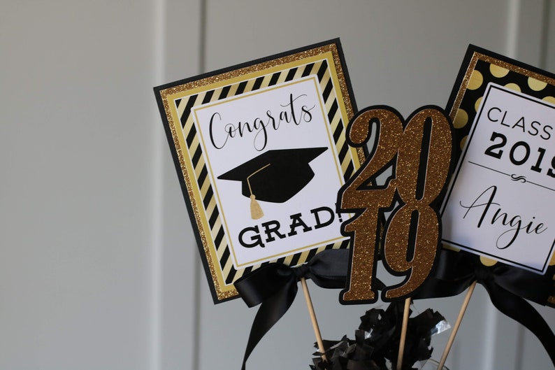 Puede incluir: Decoraciones para una fiesta de graduaci&oacute;n con acentos negros y dorados. Las decoraciones incluyen un letrero que dice "Congrats Grad!" con un gorro de graduaci&oacute;n, un letrero que dice "Class of 2019 Angie", y un gran centro de mesa de purpurina dorada "2019".