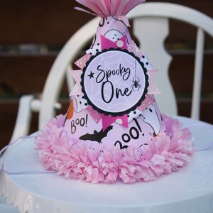 May include: A pink Halloween party hat with a decorative pink tissue paper trim. The hat features a "Spooky One" design with a spider, bats, and ghosts. The hat is on a white cake stand.