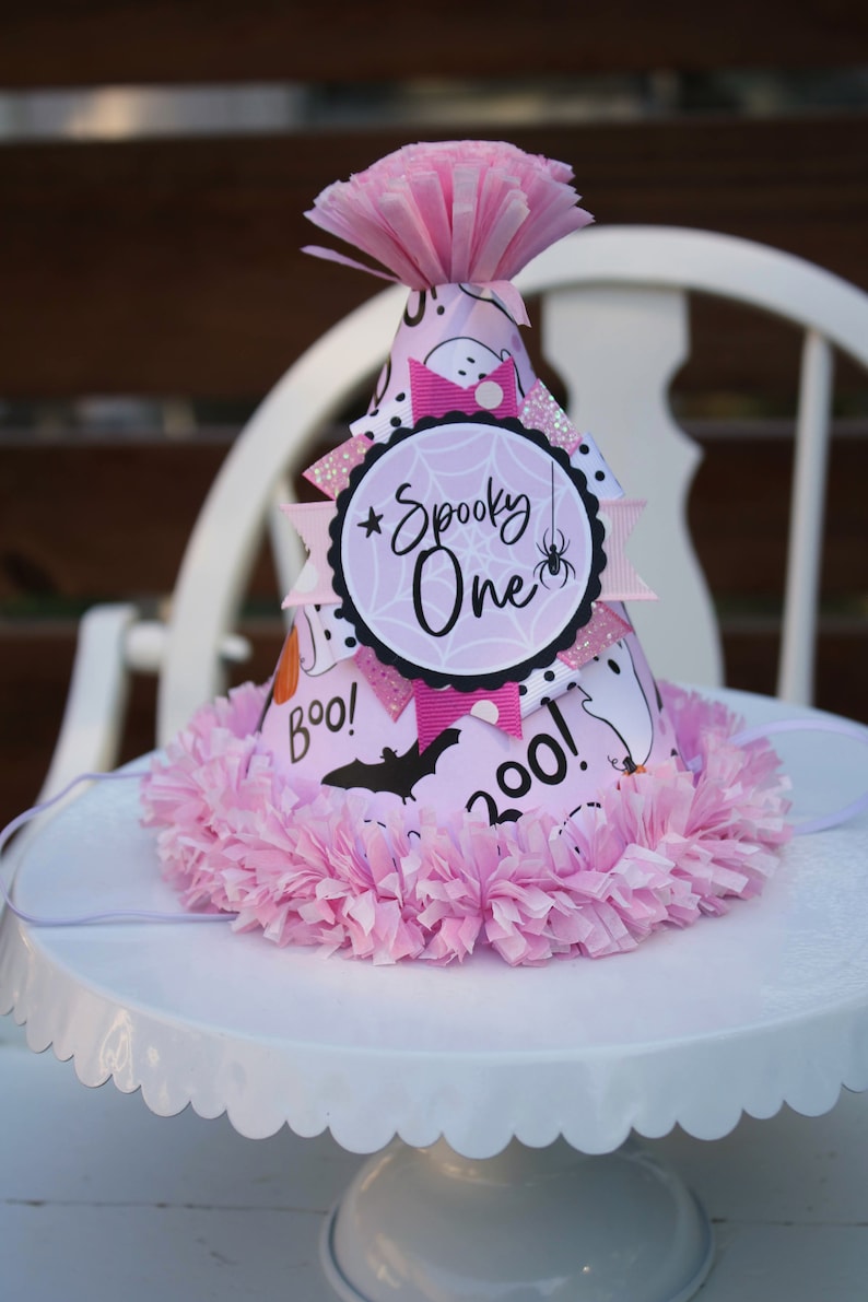 May include: A pink Halloween party hat with a decorative pink tissue paper trim. The hat features a "Spooky One" design with a spider, bats, and ghosts. The hat is on a white cake stand.