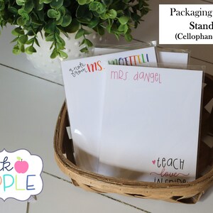 May include: Three white note cards with colorful text, including "a note from MS. ETTELI", "MRS. dangel", and "teach love inside". The cards are in a brown wicker basket with a cellophane sleeve. The text "pink APPLE" is visible in the lower left corner of the image.