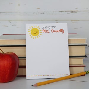 Puede incluir: Un bloc de notas blanco con un sol amarillo sonriente y el texto "A note from Mrs. Connelly" en la parte superior. Una manzana roja, un l&aacute;piz amarillo y una pila de libros est&aacute;n sobre una superficie blanca.