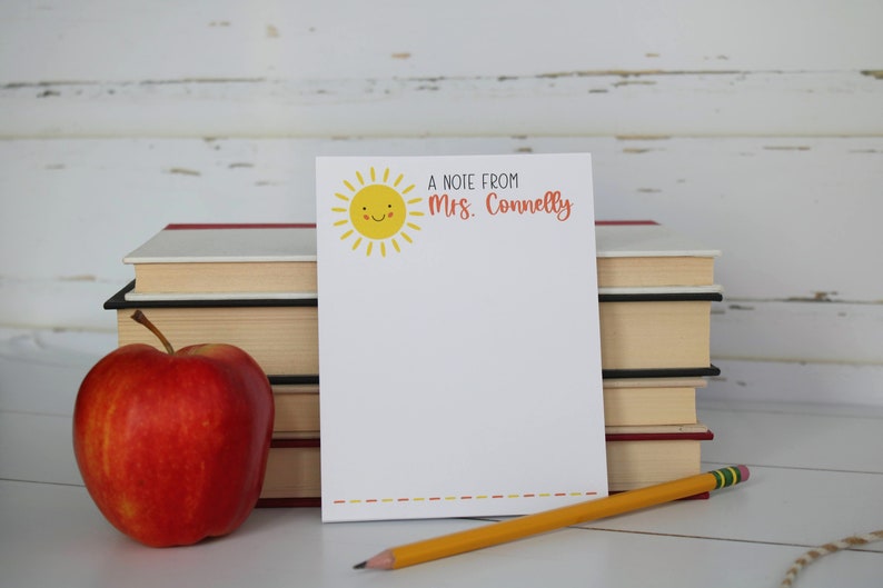 Puede incluir: Un bloc de notas blanco con un sol amarillo sonriente y el texto "A note from Mrs. Connelly" en la parte superior. Una manzana roja, un l&aacute;piz amarillo y una pila de libros est&aacute;n sobre una superficie blanca.