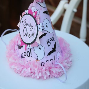 May include: A pink Halloween-themed party hat with a decorative pink pom-pom trim. The hat features ghost and bat graphics, the word "Boo!", and a circular emblem that says "Spooky One". A white string is attached.