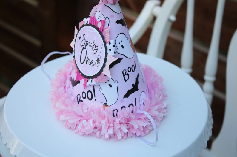 May include: A pink Halloween-themed party hat with a decorative pink pom-pom trim. The hat features ghost and bat graphics, the word "Boo!", and a circular emblem that says "Spooky One". A white string is attached.