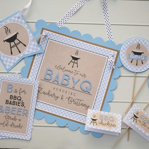 May include: Blue and brown baby shower decorations with a barbecue theme. The decorations include a banner, a sign, and several small signs with text such as "Welcome to our BabyQ", "B is for BBQ, Babies, & Beer", "BBQ chicken", and "Corn on the cob".