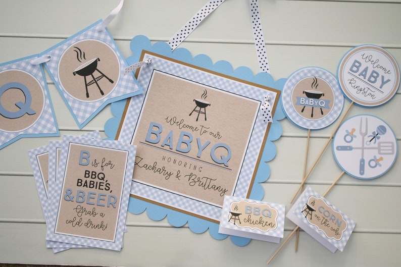 May include: Blue and brown baby shower decorations with a barbecue theme. The decorations include a banner, a sign, and several small signs with text such as "Welcome to our BabyQ", "B is for BBQ, Babies, & Beer", "BBQ chicken", and "Corn on the cob".