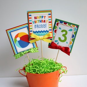 May include: A colorful centerpiece for a birthday party with a beach theme. The centerpiece features three cards with the text "Happy Birthday Brody!" and the number "3" on them. The cards are attached to wooden skewers and placed in a small orange bucket filled with green grass.