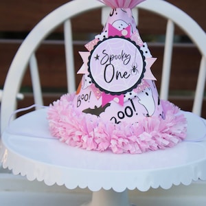 May include: A pink Halloween party hat with a ruffled base and a paper flower on top. The hat is decorated with ghosts, bats, and the words "Spooky One". It sits on a white cake stand.