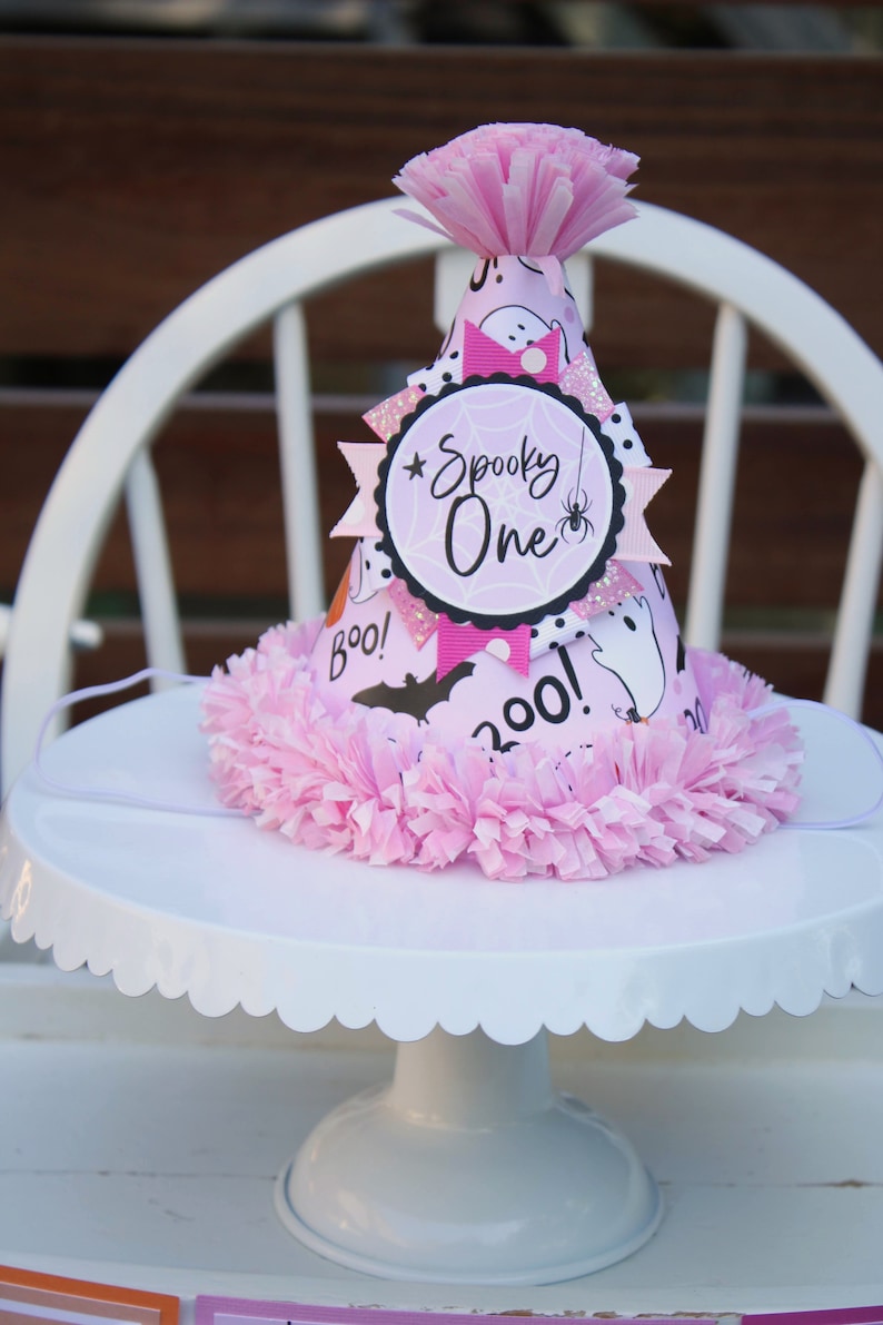 May include: A pink Halloween party hat with a ruffled base and a paper flower on top. The hat is decorated with ghosts, bats, and the words "Spooky One". It sits on a white cake stand.