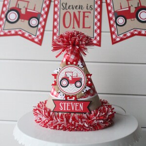 May include: Red and white checkered birthday party hat with a red tractor design and the name "Steven" on a banner. The hat is decorated with a red and white pom-pom.
