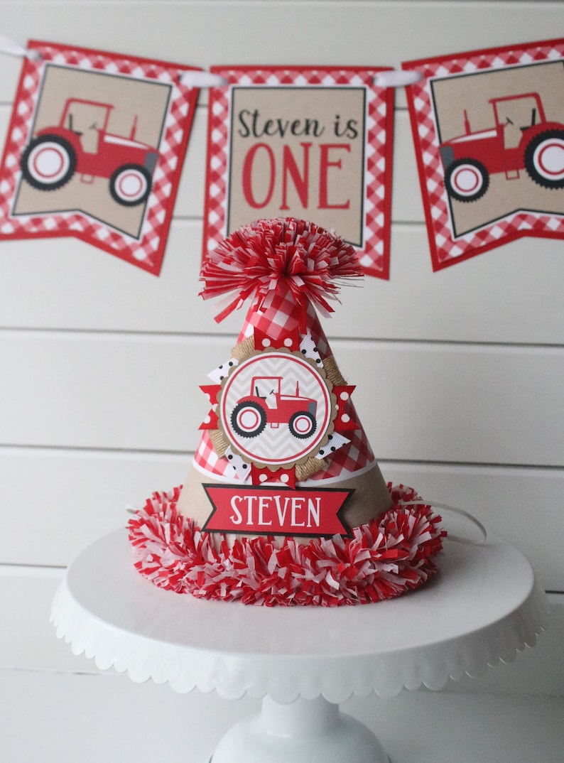 May include: Red and white checkered birthday party hat with a red tractor design and the name "Steven" on a banner. The hat is decorated with a red and white pom-pom.