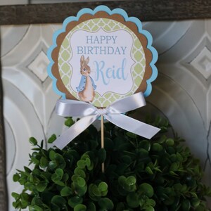 K&ouml;nnte beinhalten: Geburtstagskuchenaufsatz mit Peter Rabbit-Motiv. Der Aufsatz hat einen gewellten Rand mit blauen und braunen Akzenten und den Text "HAPPY BIRTHDAY Reid". Eine wei&szlig;e Schleife ist an dem Holzstab befestigt.