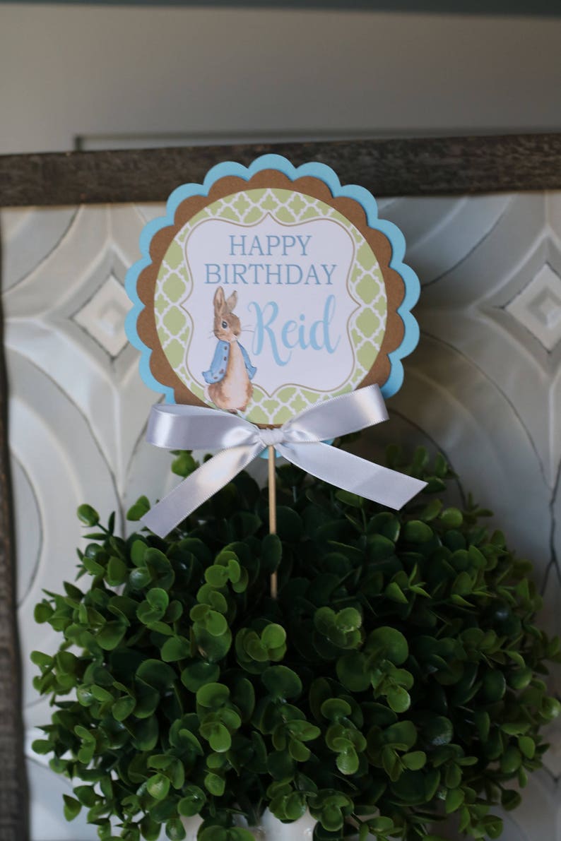 K&ouml;nnte beinhalten: Geburtstagskuchenaufsatz mit Peter Rabbit-Motiv. Der Aufsatz hat einen gewellten Rand mit blauen und braunen Akzenten und den Text "HAPPY BIRTHDAY Reid". Eine wei&szlig;e Schleife ist an dem Holzstab befestigt.